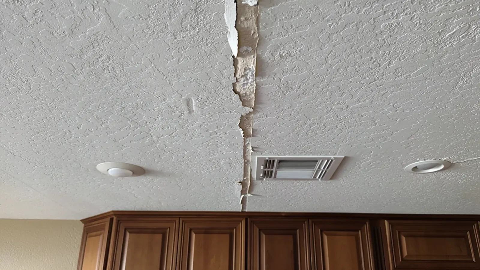 A crack running through a textured kitchen ceiling beside a white rectangular ceiling air register, the paint and joint tape split open along its length, above stained wood upper cabinets