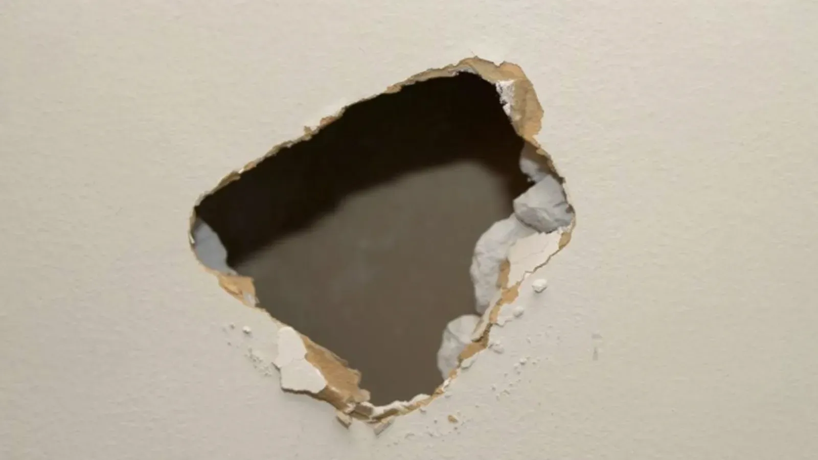 Large irregular hole broken through a painted interior drywall wall, torn paper facing and crumbling gypsum chunks around the jagged edges exposing the dark cavity behind