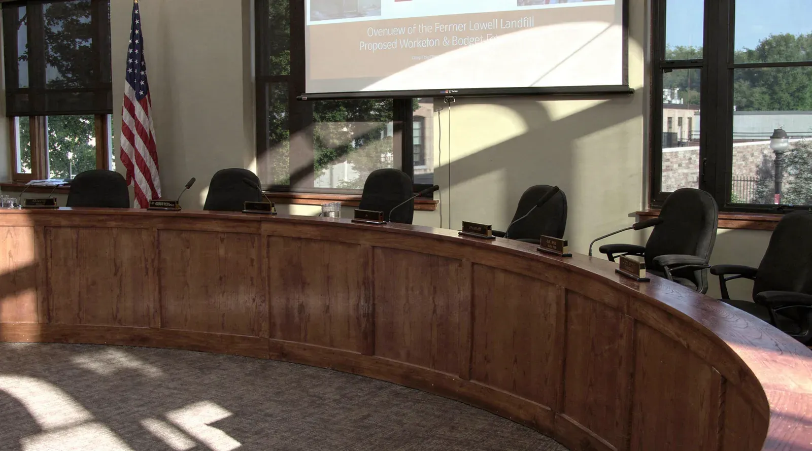Lowell municipal council chamber with a curved oak dais, table microphones, the United States flag, and plaster-finished walls beneath tall windows