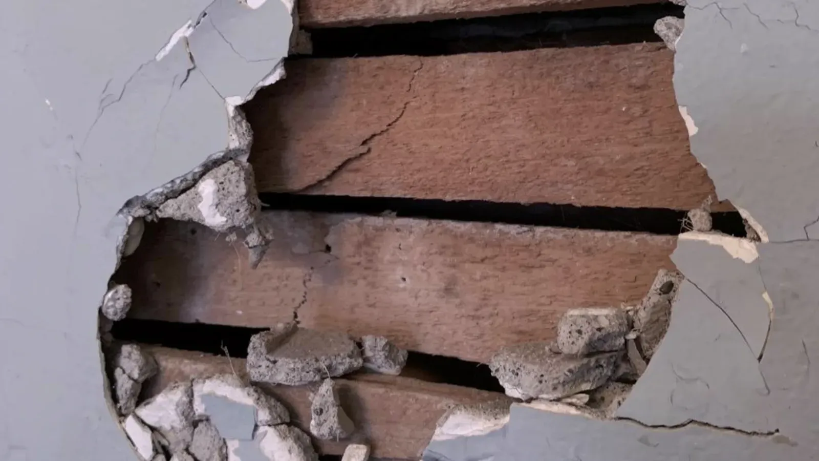 Hole broken through a painted plaster wall, exposing the horizontal wood lath behind it and the crumbling plaster keys around the opening